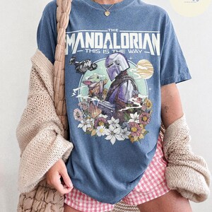 K&ouml;nnte beinhalten: Blaues Jeans-T-Shirt mit einer Grafik von The Mandalorian und Grogu, umgeben von Blumen und einem Planeten. Der Text "THE MANDALORIAN" und "THIS IS THE WAY" sind &uuml;ber der Grafik aufgedruckt. Das Shirt ist Teil der "COMFORT COLORS"-Kollektion.