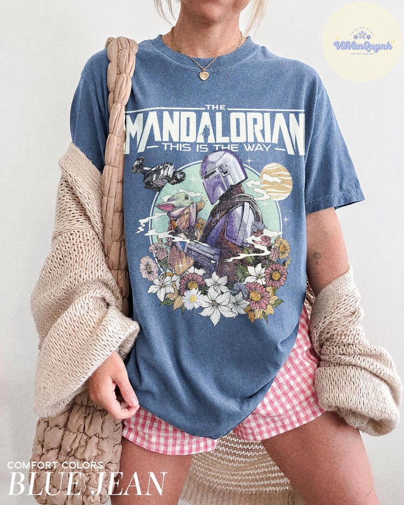 K&ouml;nnte beinhalten: Blaues Jeans-T-Shirt mit einer Grafik von The Mandalorian und Grogu, umgeben von Blumen und einem Planeten. Der Text "THE MANDALORIAN" und "THIS IS THE WAY" sind &uuml;ber der Grafik aufgedruckt. Das Shirt ist Teil der "COMFORT COLORS"-Kollektion.