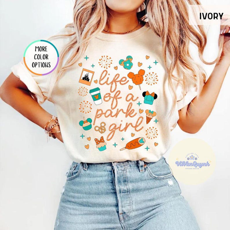 May include: Ivory-colored t-shirt with the text "life of a park girl" in orange script. The shirt features various cartoon graphics, including a coffee cup, Mickey Mouse ears, and a cupcake. The shirt is paired with blue denim jeans.