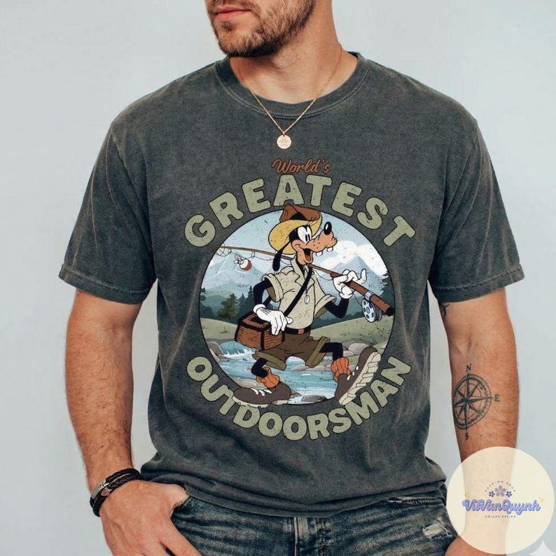 Pu&ograve; includere: T-shirt grigio scuro con un cartone animato di Pippo come pescatore, con la scritta "World's Greatest Outdoorsman" in un design circolare. La grafica include una canna da pesca, una scatola per attrezzi e un paesaggio montano.