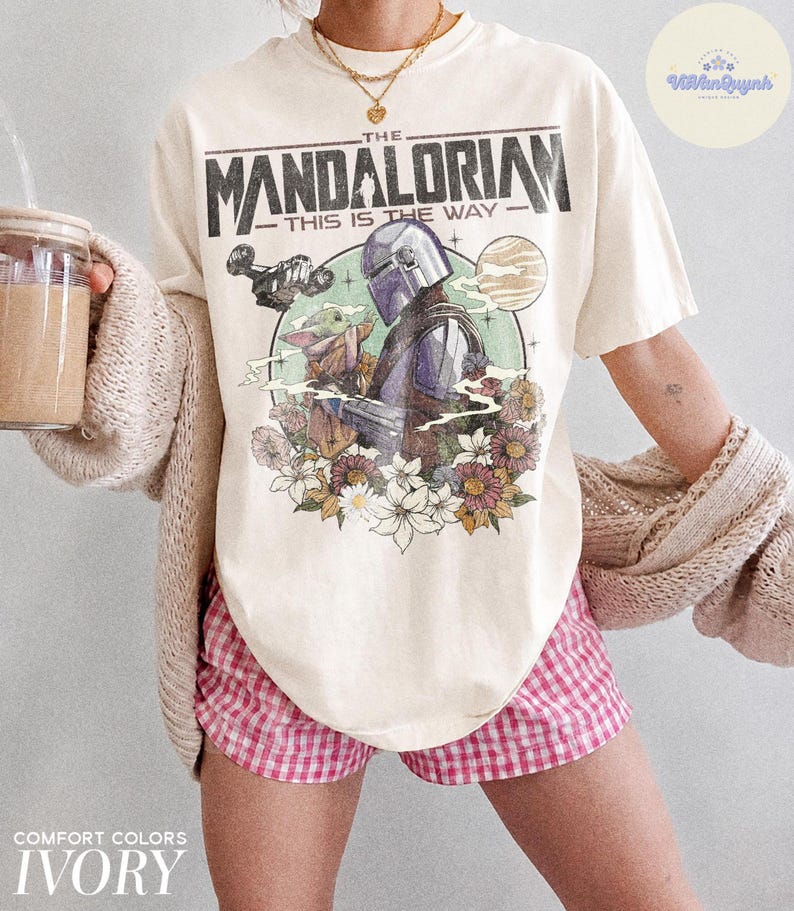 K&ouml;nnte beinhalten: Cremefarbenes T-Shirt mit einem Aufdruck von The Mandalorian und Grogu, umgeben von Blumen. Der Text "THE MANDALORIAN - THIS IS THE WAY" ist oben aufgedruckt. Das Shirt geh&ouml;rt zur Comfort Colors Kollektion.