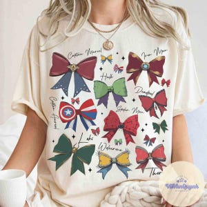 May include: Cream-colored t-shirt featuring colorful bow designs representing Marvel characters like Captain Marvel, Iron Man, and Spider-Man. Each bow has a unique color scheme and is labeled with the character's name. The shirt has a relaxed fit.