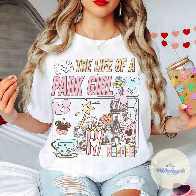 May include: White t-shirt with the text "THE LIFE OF A PARK GIRL" in pink and beige. The shirt features cartoon illustrations of a teacup, popcorn, a castle, and Mickey Mouse icons. A clear glass with a straw is held in the hand.