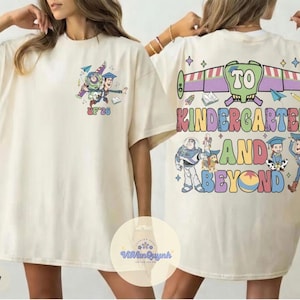 May include: Ivory-colored t-shirt featuring a colorful graphic of Buzz Lightyear and Woody from Toy Story, with the text "UF '26" on the front. The back of the shirt displays the phrase "TO KINDERGARTEN AND BEYOND" with additional Toy Story characters.