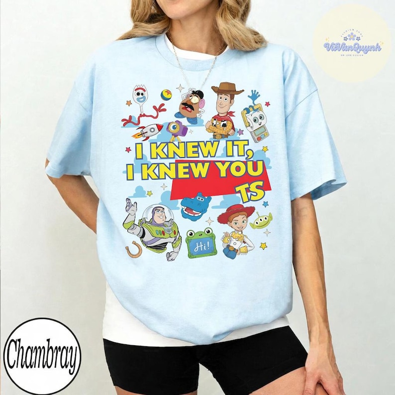 Op de afbeelding: Een lichtblauw t-shirt met een kleurrijke grafische print van diverse Toy Story-personages, waaronder Buzz Lightyear, Woody en Mr. Potato Head. De tekst luidt I KNEW IT, I KNEW YOU TS, met een rood rechthoekig blok achter het woord TS.