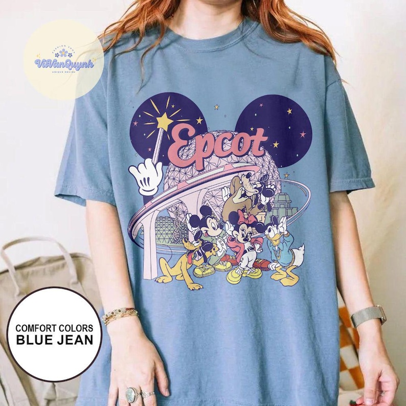 May include: Blue jean colored t-shirt featuring a graphic of Disney characters, including Mickey and Minnie Mouse, Goofy, and Donald Duck. The word "Epcot" is printed in pink script. The shirt has a round neck and short sleeves.