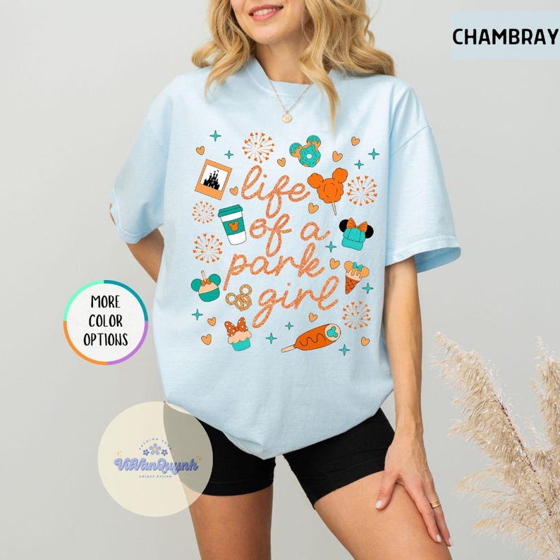 May include: Light blue t-shirt with the text "life of a park girl" and various graphic icons. The icons include a castle, coffee cup, Mickey Mouse ears, and a cupcake. The shirt also has the text "MORE COLOR OPTIONS" on the left side.