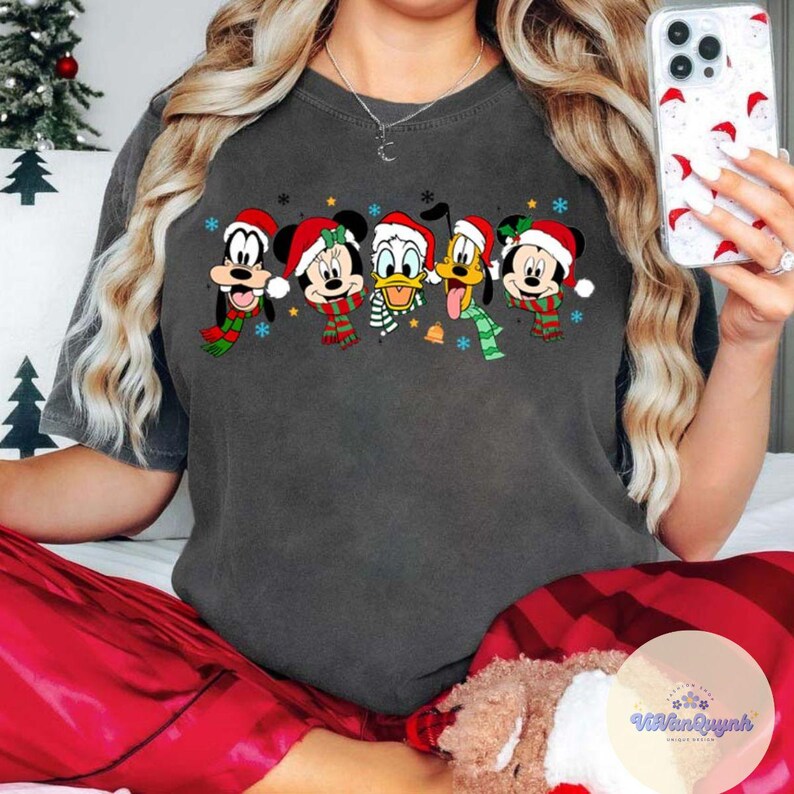 May include: Dark gray t-shirt featuring a festive design with cartoon characters wearing Santa hats and scarves. The characters include Mickey Mouse, Minnie Mouse, Donald Duck, Goofy, and Pluto, with snowflakes and bells. A Christmas-themed apparel.