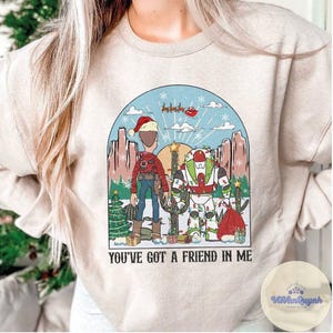 May include: Beige crewneck sweatshirt with a colorful graphic featuring two cartoon characters in a desert scene with a Christmas theme. The text "YOU'VE GOT A FRIEND IN ME" is printed below the graphic. A Christmas tree, presents, and Santa's sleigh are also visible.