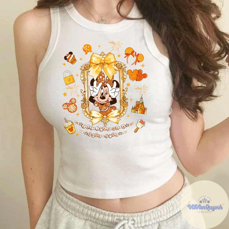May include: White tank top featuring a Minnie Mouse graphic in a gold frame with bows and other themed illustrations. The design includes a castle, ice cream, and a coffee cup. The tank top is sleeveless and has a round neckline.
