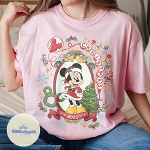 May include: Pink t-shirt featuring a festive Minnie Mouse design. The graphic includes Minnie in a Santa outfit, a Christmas tree, and the words "Christmas" and "Very Merry" in a decorative frame. The year "2025" is also displayed.