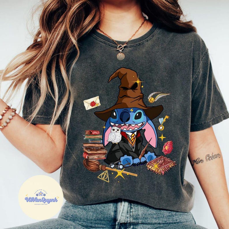 May include: A dark grey t-shirt featuring a blue Stitch character dressed as a wizard, wearing a brown pointed hat and holding a wand. The design also includes a white owl, books, a broom, and a golden snitch.