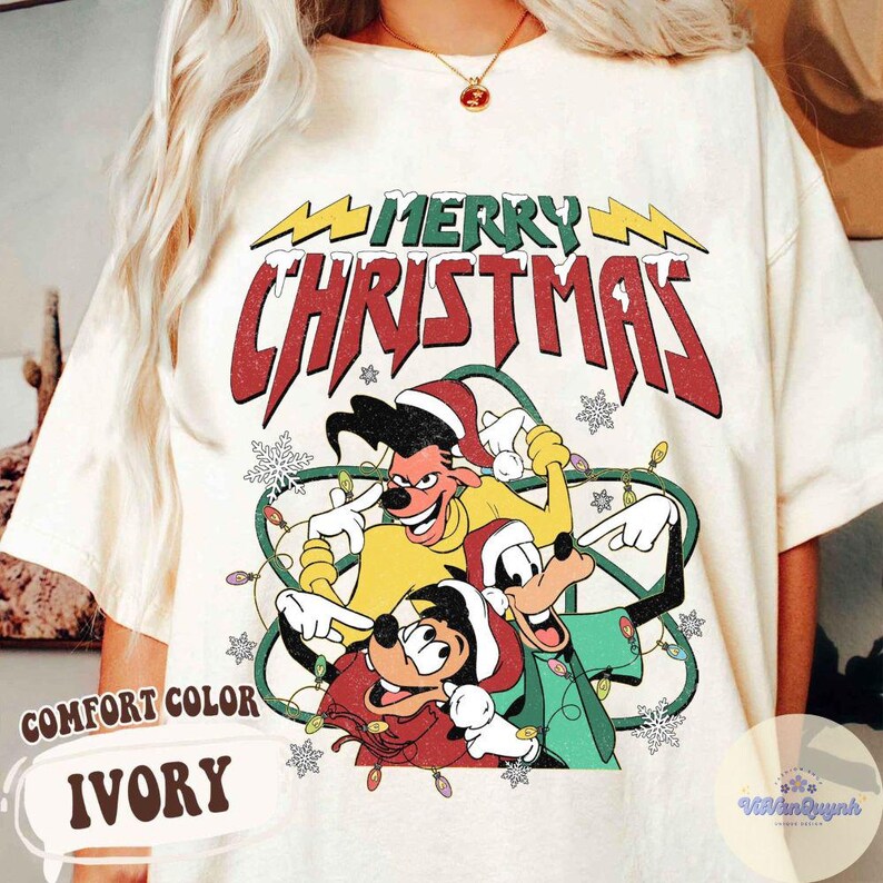 May include: Ivory Comfort Color t-shirt with a graphic of cartoon characters and the text "MERRY CHRISTMAS" in red and green. The design includes snowflakes and Christmas lights.