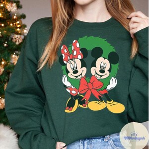 Op de afbeelding: Donkergroene sweatshirt met een feestelijke afbeelding van Mickey en Minnie Mouse in een groene krans, versierd met een rode strik. Minnie draagt een rode strik met stippen. Het ontwerp heeft gele, rode, witte en zwarte accenten.