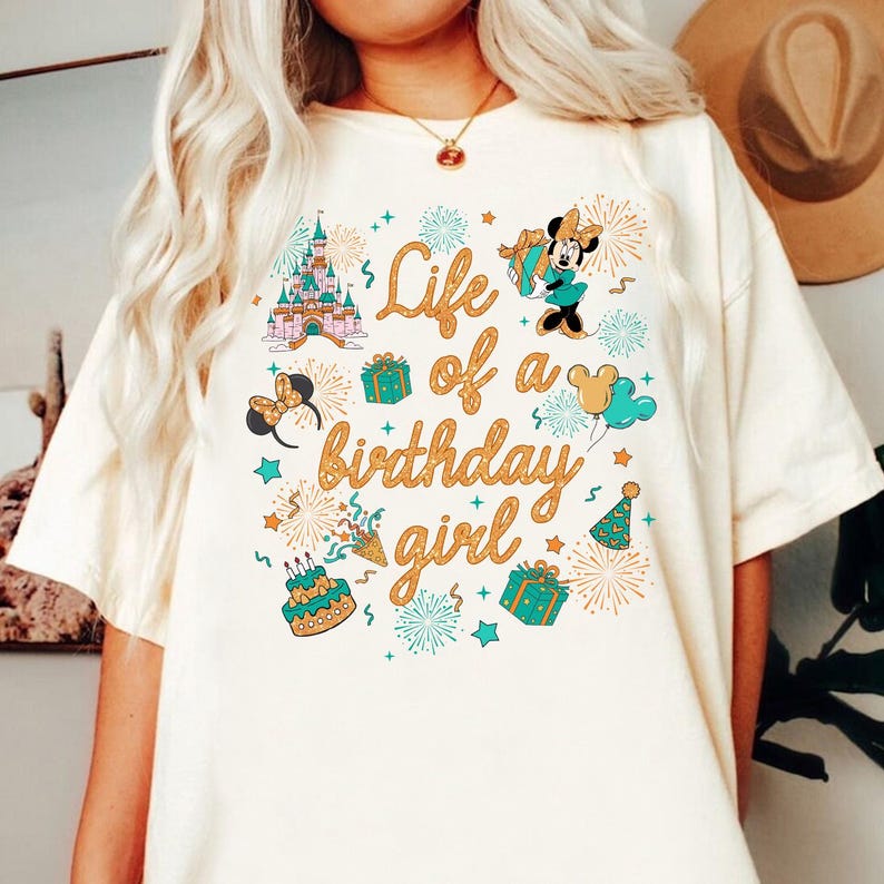 Pu&ograve; includere: T-shirt color crema con un design grafico colorato. Il design include un castello, Minnie Mouse, regali, fuochi d'artificio e la scritta "Life of a birthday girl" in oro. Elementi aggiuntivi: torta di compleanno e cappello da festa.