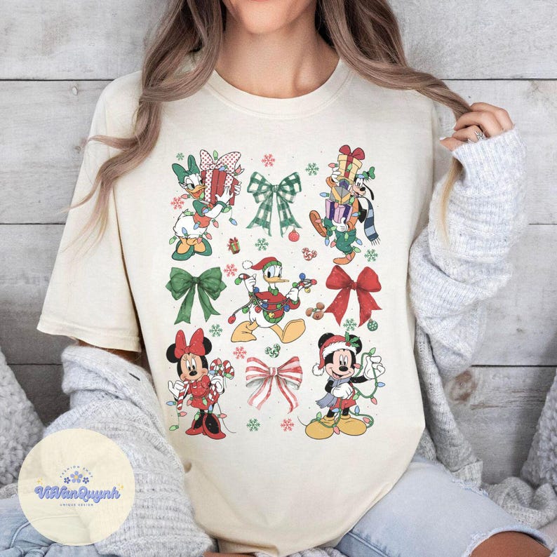 May include: Cream-colored t-shirt featuring a festive design with Disney characters and Christmas bows. The graphic includes Mickey Mouse, Minnie Mouse, Donald Duck, and Goofy, each adorned with holiday lights and gifts. Snowflakes and bows complete the seasonal theme.