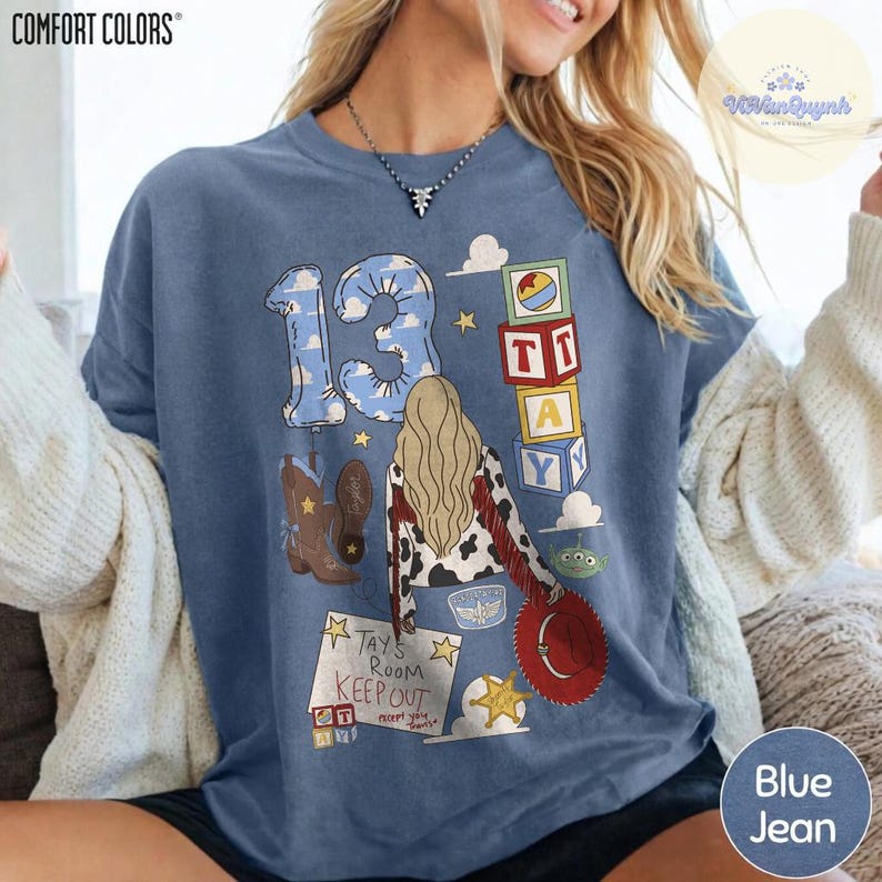 May include: A blue short-sleeve t-shirt featuring a graphic print with the number 13, a person with long blonde hair, cowboy boots, toy blocks, and a sign that reads "TAY'S ROOM KEEP OUT". The shirt has a crew neckline and a relaxed fit.