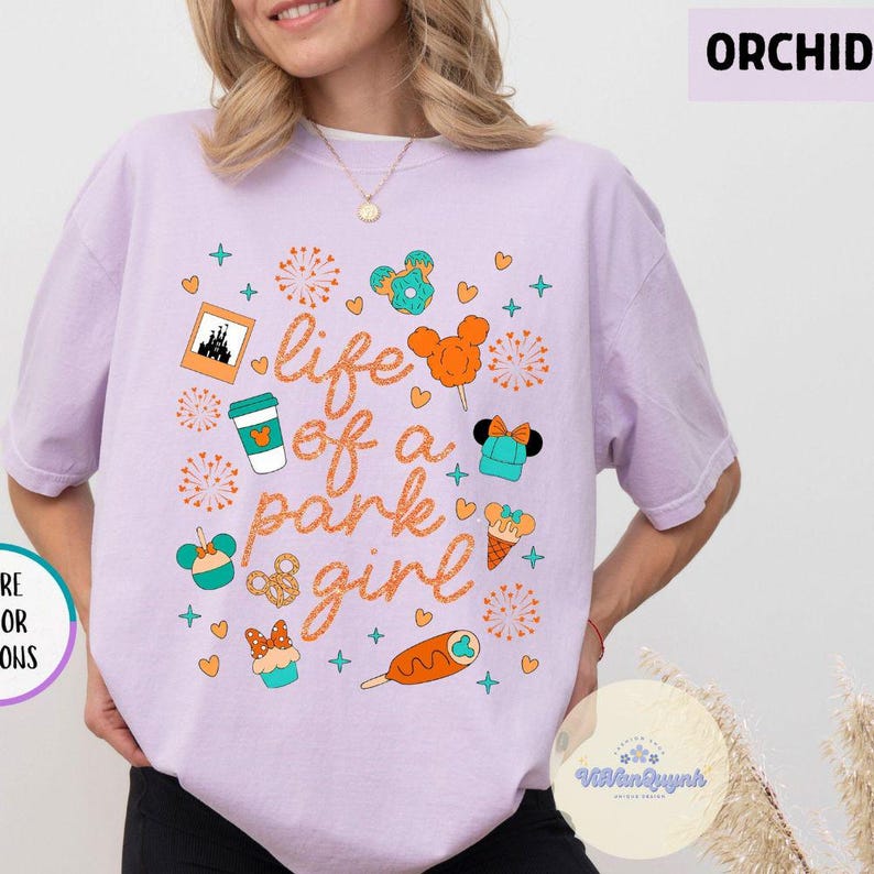 May include: Orchid-colored t-shirt with a graphic design featuring theme park icons and the text "life of a park girl" in orange script. The design includes Mickey Mouse-shaped treats, a castle, and fireworks.