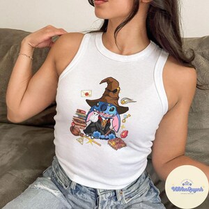 May include: A white tank top featuring a blue Stitch character dressed as a wizard, wearing a brown hat and holding a book. The design includes other wizarding items such as a wand, a book, and a potion bottle. The tank top has a white background and the Stitch character is centrally positioned.