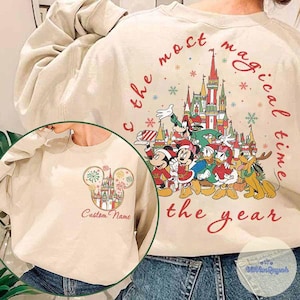 Puede incluir: Sudadera beige con un gr&aacute;fico colorido de personajes de Disney frente a un castillo, con el texto "the most magical time of the year". Una imagen m&aacute;s peque&ntilde;a muestra una cabeza de Mickey Mouse con un castillo y las palabras "Custom Name".
