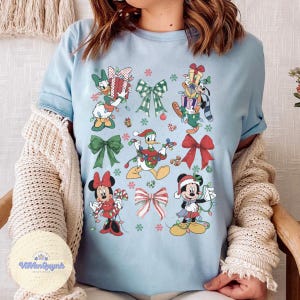May include: Light blue t-shirt featuring Christmas-themed Disney characters, including Mickey and Minnie Mouse, Donald Duck, and Goofy, adorned with festive lights and bows. Snowflakes and gift boxes are also included in the design.