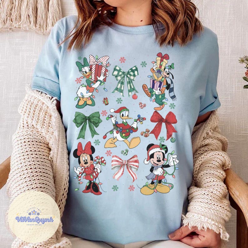 May include: Light blue t-shirt featuring Christmas-themed Disney characters, including Mickey and Minnie Mouse, Donald Duck, and Goofy, adorned with festive lights and bows. Snowflakes and gift boxes are also included in the design.