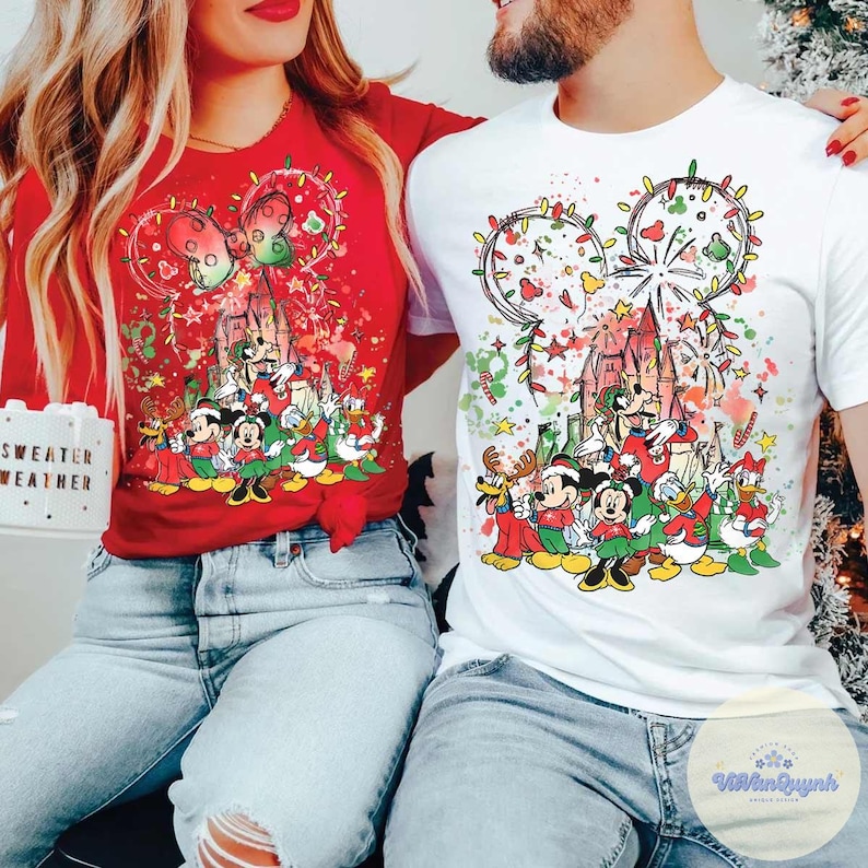 May include: Red and white Christmas-themed t-shirts featuring a Disney character design. The design includes Mickey Mouse, Minnie Mouse, Donald Duck, and other characters in festive attire. The shirts have a castle and fireworks design.