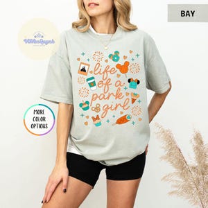 May include: A light gray t-shirt with the text "life of a park girl" in orange script. The shirt features colorful graphics of theme park icons, including a castle, Mickey Mouse balloons, and snacks. The shirt is a relaxed fit.