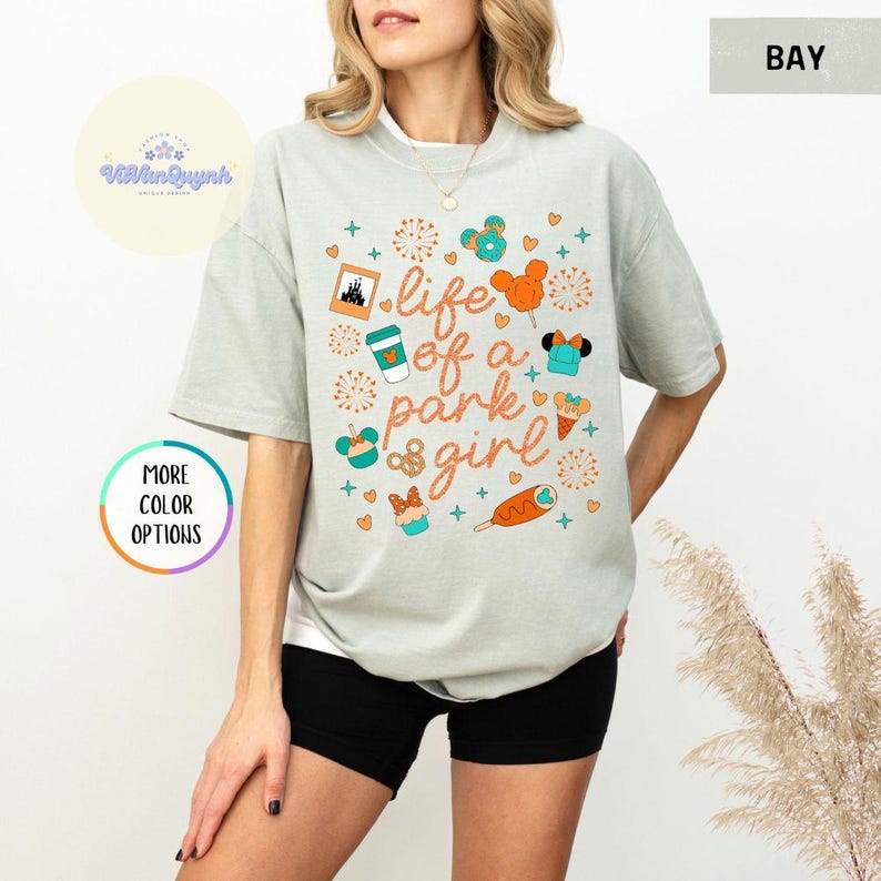 May include: A light gray t-shirt with the text "life of a park girl" in orange script. The shirt features colorful graphics of theme park icons, including a castle, Mickey Mouse balloons, and snacks. The shirt is a relaxed fit.