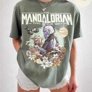 K&ouml;nnte beinhalten: Moosgr&uuml;nes T-Shirt mit einer Grafik von The Mandalorian und Grogu, umgeben von Blumen. Der Text "THE MANDALORIAN - THIS IS THE WAY" ist oben aufgedruckt. Das Shirt besteht aus Comfort Colors Material.