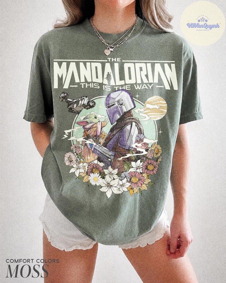 K&ouml;nnte beinhalten: Moosgr&uuml;nes T-Shirt mit einer Grafik von The Mandalorian und Grogu, umgeben von Blumen. Der Text "THE MANDALORIAN - THIS IS THE WAY" ist oben aufgedruckt. Das Shirt besteht aus Comfort Colors Material.
