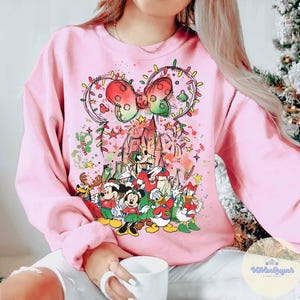 May include: Pink sweatshirt featuring a festive Disney design. The graphic includes Mickey and Minnie Mouse, Donald Duck, and Goofy, with a castle, Christmas lights, and a bow. The design is colorful and whimsical, perfect for the holiday season.