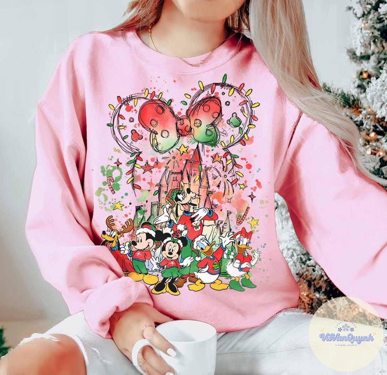 May include: Pink sweatshirt featuring a festive Disney design. The graphic includes Mickey and Minnie Mouse, Donald Duck, and Goofy, with a castle, Christmas lights, and a bow. The design is colorful and whimsical, perfect for the holiday season.