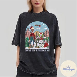 May include: Black t-shirt featuring a graphic of two cartoon characters in a winter scene with a Christmas tree and cactus. The text "YOU'VE GOT A FRIEND IN ME" is printed below the graphic. The shirt has short sleeves and a crew neck.