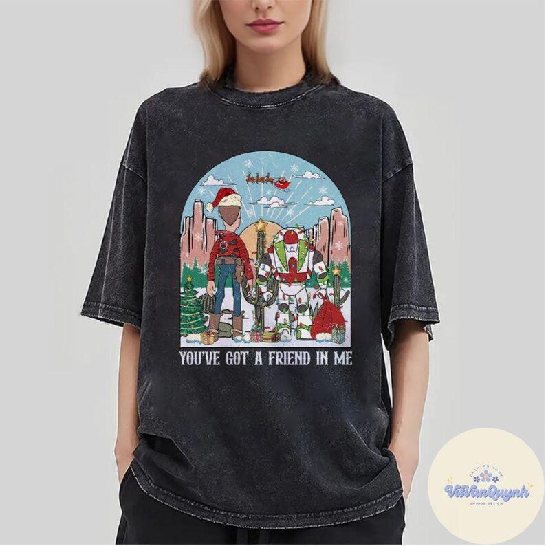 May include: Black t-shirt featuring a graphic of two cartoon characters in a winter scene with a Christmas tree and cactus. The text "YOU'VE GOT A FRIEND IN ME" is printed below the graphic. The shirt has short sleeves and a crew neck.