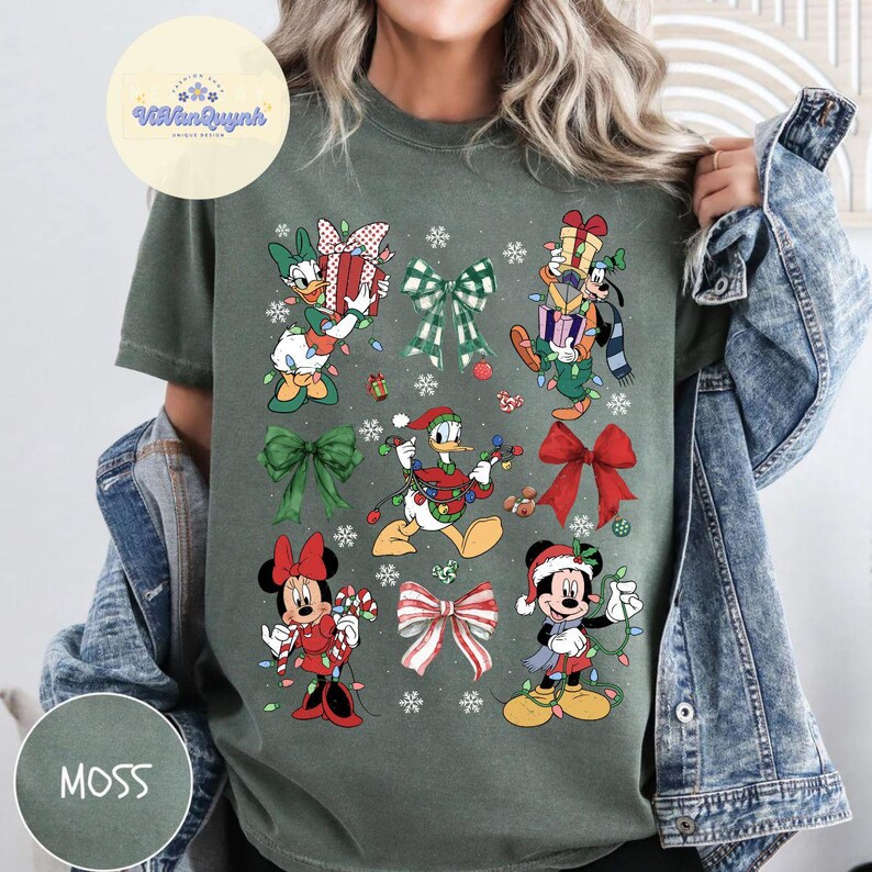 May include: Moss green t-shirt featuring Disney characters in Christmas attire. Donald Duck, Mickey and Minnie Mouse, and Goofy are depicted with gifts, lights, and bows. The shirt has the word "MOSS" printed on the bottom left.