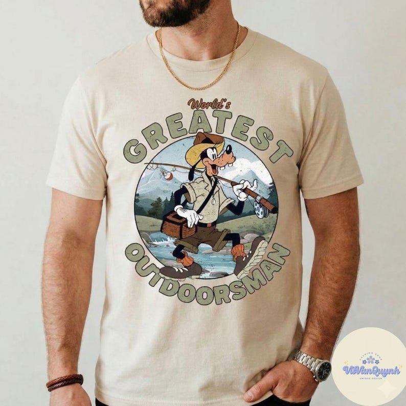 Pu&ograve; includere: T-shirt beige con un'illustrazione a cartoni animati di Pippo come un uomo all'aperto, con una canna da pesca e una scatola per attrezzi. Il disegno &egrave; in una cornice circolare con la scritta "World's Greatest Outdoorsman".
