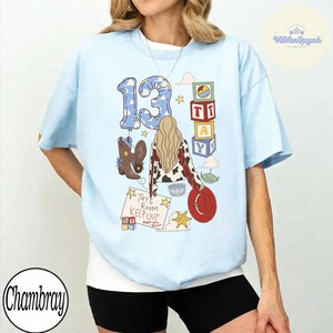 May include: A light blue t-shirt featuring a graphic print with the number 13, a person with long blonde hair wearing a cow-print jacket, brown cowboy boots, a red hat, colorful alphabet blocks, and a sign that reads Tay's Room Keep Out.