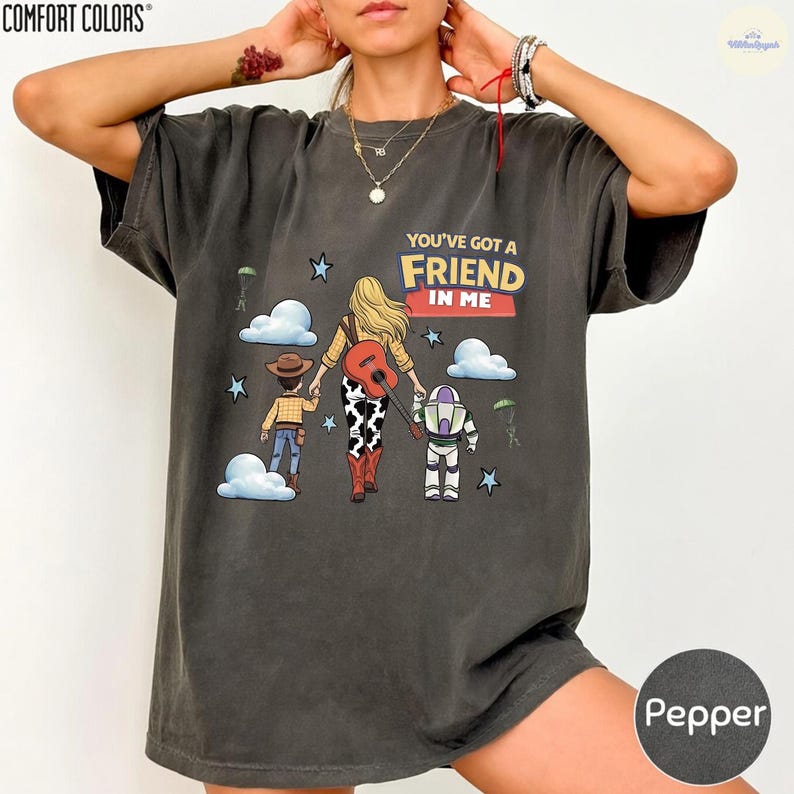 Op de afbeelding: Een antracietgrijs t-shirt met korte mouwen en een afbeelding van een persoon met blond haar die hand in hand loopt met een cowboy en een space ranger. De tekst luidt YOU'VE GOT A FRIEND IN ME, met wolken, sterren en een gitaar.