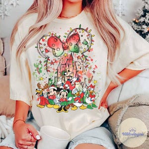 May include: Cream-colored t-shirt featuring a festive Disney design. The graphic includes Mickey Mouse ears adorned with Christmas lights, a castle, and various Disney characters in holiday attire. The design incorporates red, green, and yellow accents.