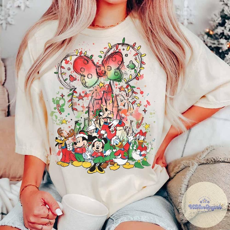 May include: Cream-colored t-shirt featuring a festive Disney design. The graphic includes Mickey Mouse ears adorned with Christmas lights, a castle, and various Disney characters in holiday attire. The design incorporates red, green, and yellow accents.