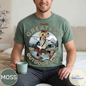 Pu&ograve; includere: T-shirt verde muschio con grafica circolare di Pippo pescatore, con la scritta "World's Greatest Outdoorsman". Il design include una canna da pesca, una scatola per esche e uno sfondo panoramico.