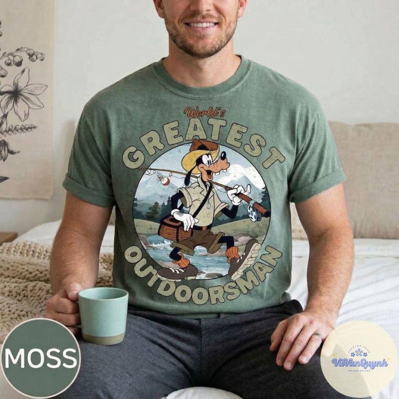 Pu&ograve; includere: T-shirt verde muschio con grafica circolare di Pippo pescatore, con la scritta "World's Greatest Outdoorsman". Il design include una canna da pesca, una scatola per esche e uno sfondo panoramico.