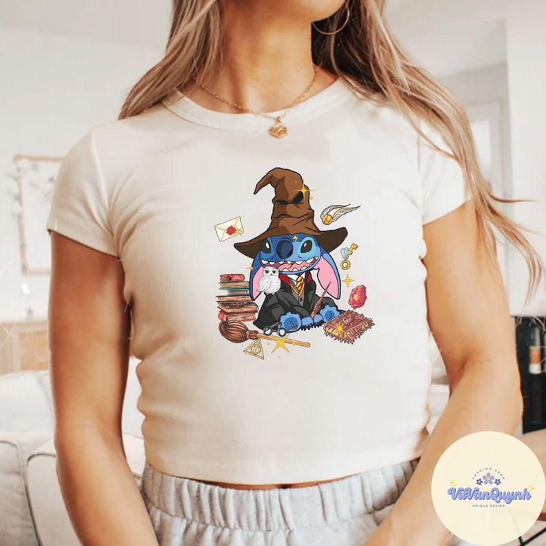May include: A white crop top featuring a blue Stitch character dressed as a witch, holding a wand and surrounded by books, a broom, and other magical items.