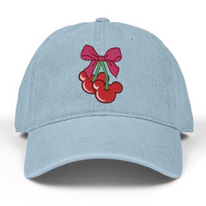 May include: Light blue baseball cap with a red and pink embroidered cherry design. The cherries have a Mickey Mouse-like shape and are tied with a pink bow. The cap has a curved brim and a button on top.