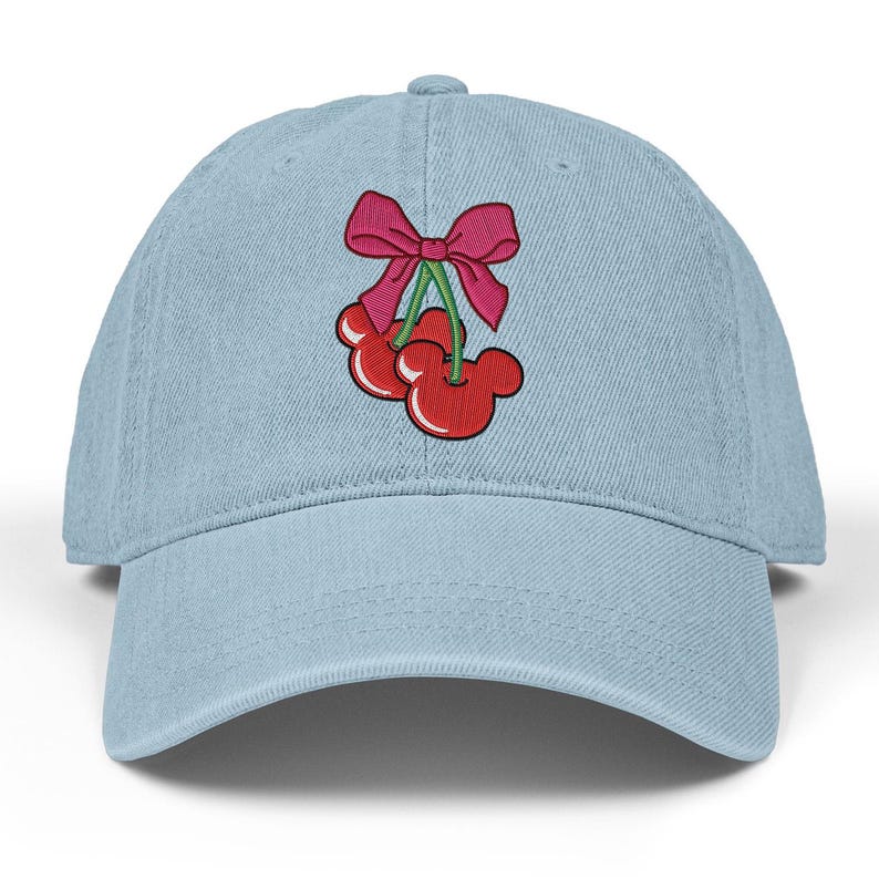 May include: Light blue baseball cap with a red and pink embroidered cherry design. The cherries have a Mickey Mouse-like shape and are tied with a pink bow. The cap has a curved brim and a button on top.