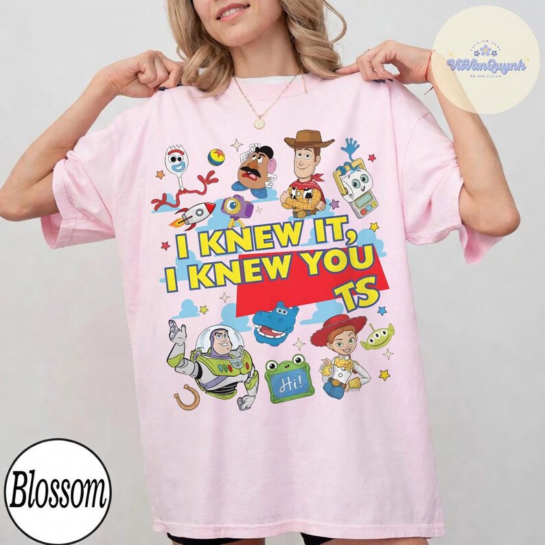 Op de afbeelding: Een lichtroze t-shirt met een kleurrijke grafische print van diverse Toy Story-personages, waaronder Woody, Buzz Lightyear, Jessie, Mr. Potato Head en Forky. De tekst I KNEW IT, I KNEW YOU TS staat in het geel en rood in het midden gedrukt.