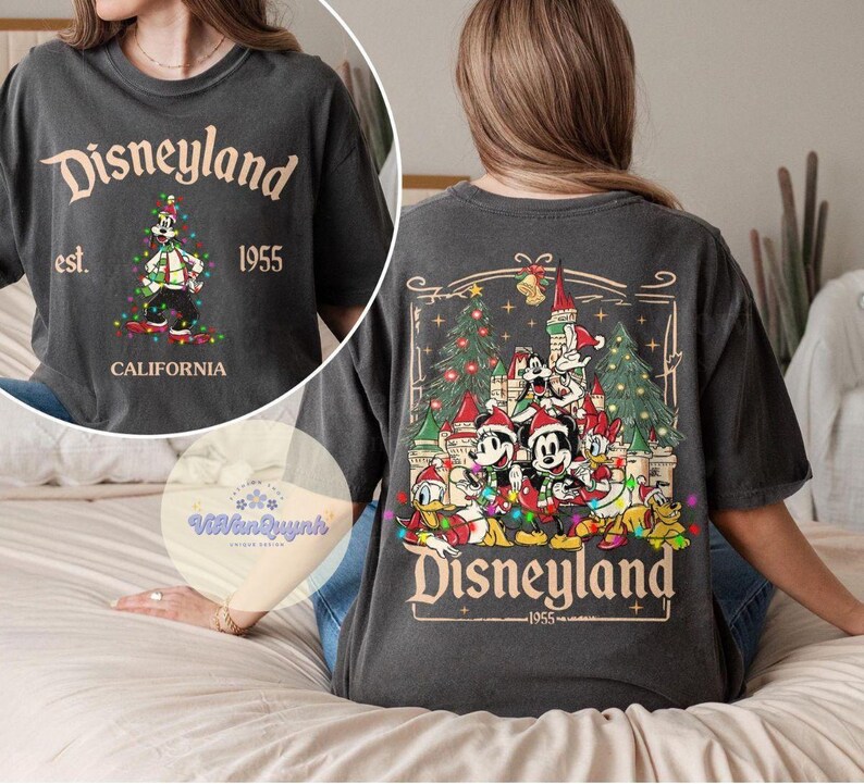 May include: Dark gray t-shirt with "Disneyland" text and a graphic of Mickey Mouse in front of a Christmas tree. The back of the shirt features a Disneyland castle scene with Mickey and friends. "Est. 1955 California" is also printed on the shirt.