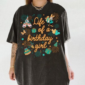 Pu&ograve; includere: T-shirt grigio scuro con la scritta "Life of a birthday girl" in arancione. La maglietta presenta grafiche colorate di un castello, Minnie Mouse, regali, fuochi d'artificio, una torta e stelle. La maglietta ha un aspetto vintage e usurato.