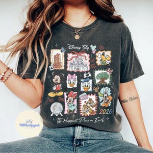 K&ouml;nnte beinhalten: Dunkelgraues T-Shirt mit einem farbenfrohen Disney Trip-Design. Das Design zeigt verschiedene Disney-Charaktere und Wahrzeichen, mit dem Text "The Happiest Place on Earth" und dem Jahr "2026".